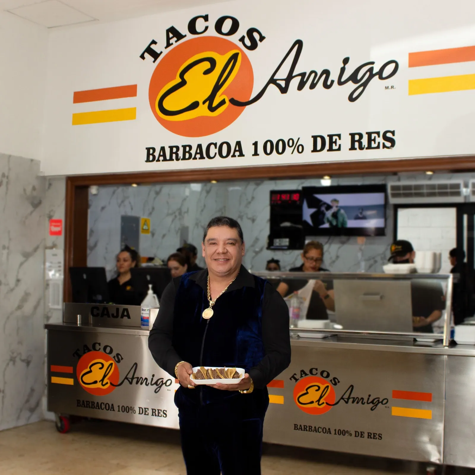 Don Guma, fundador de Tacos El Amigo, con un plato de tacos de barbacoa frente a la barra de la taquería