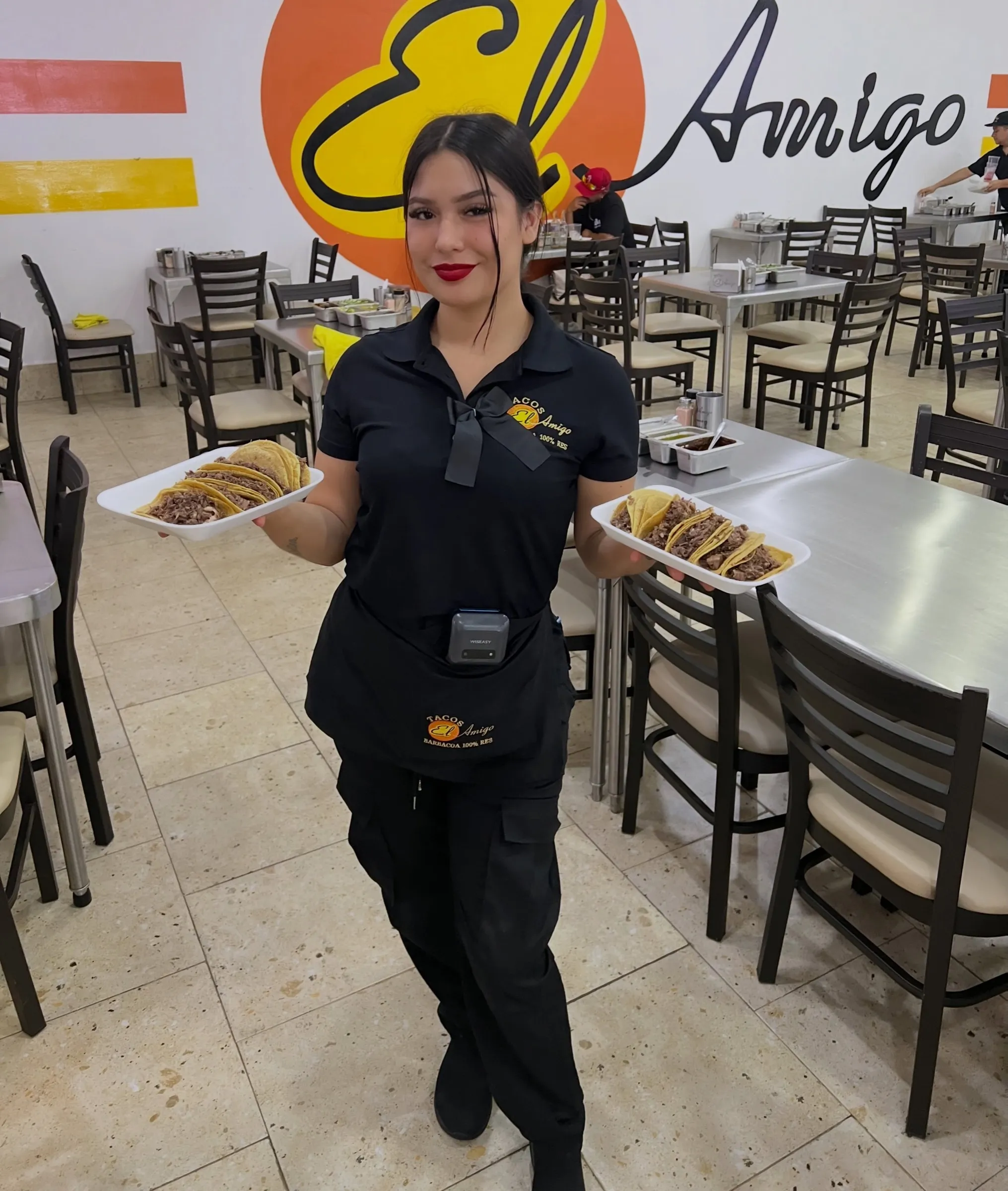Comedor de Tacos El Amigo: una mesera lleva dos platos de tacos de barbacoa frente al muro con el logo.